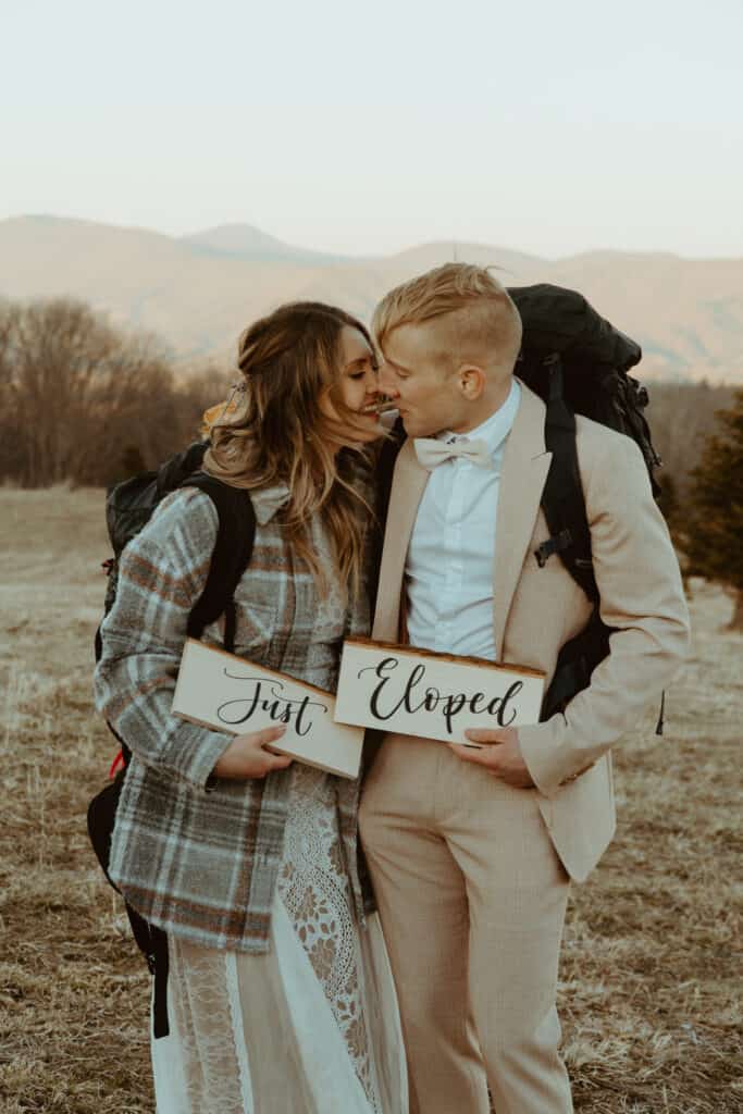 couple kissing holding just eloped signs wearing backpacks on mountain
